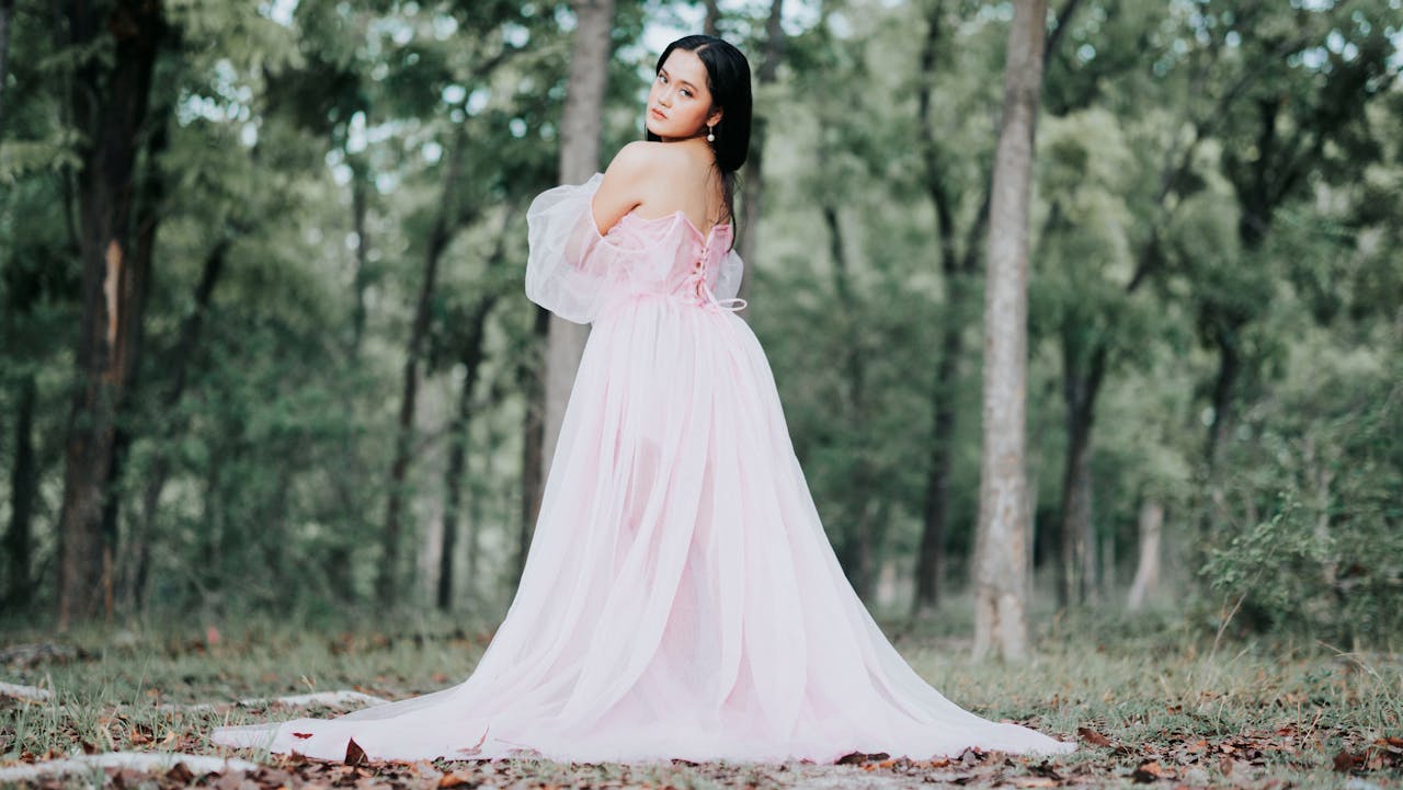 A woman in a flowing pink gown poses gracefully in a lush green forest setting.